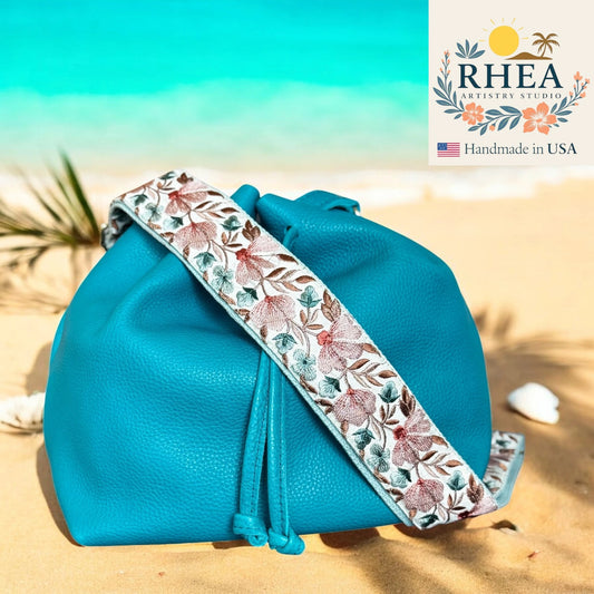 Blue bag with a floral strap on a beach, featuring the RHEA brand.