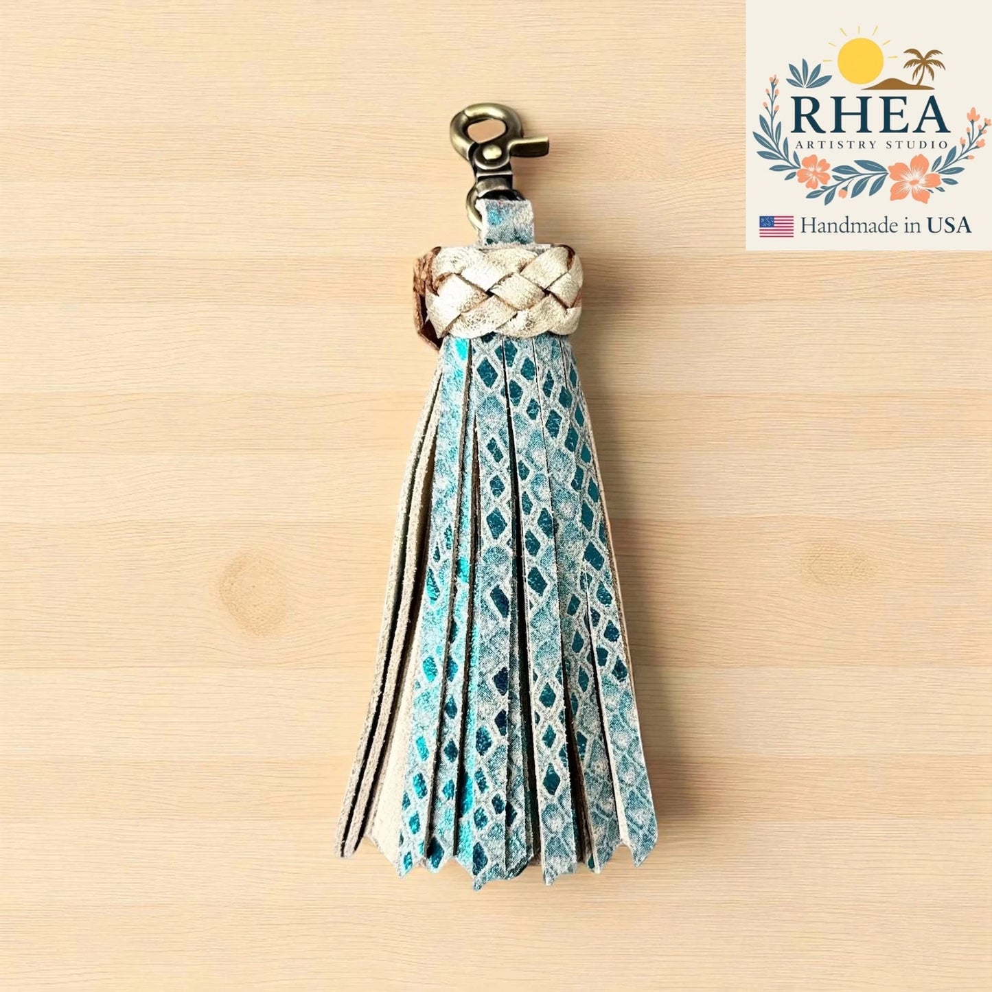 Decorative aqua silver tassel keychain with braided design on a wooden surface, featuring the RHEA Artistry Studio logo.