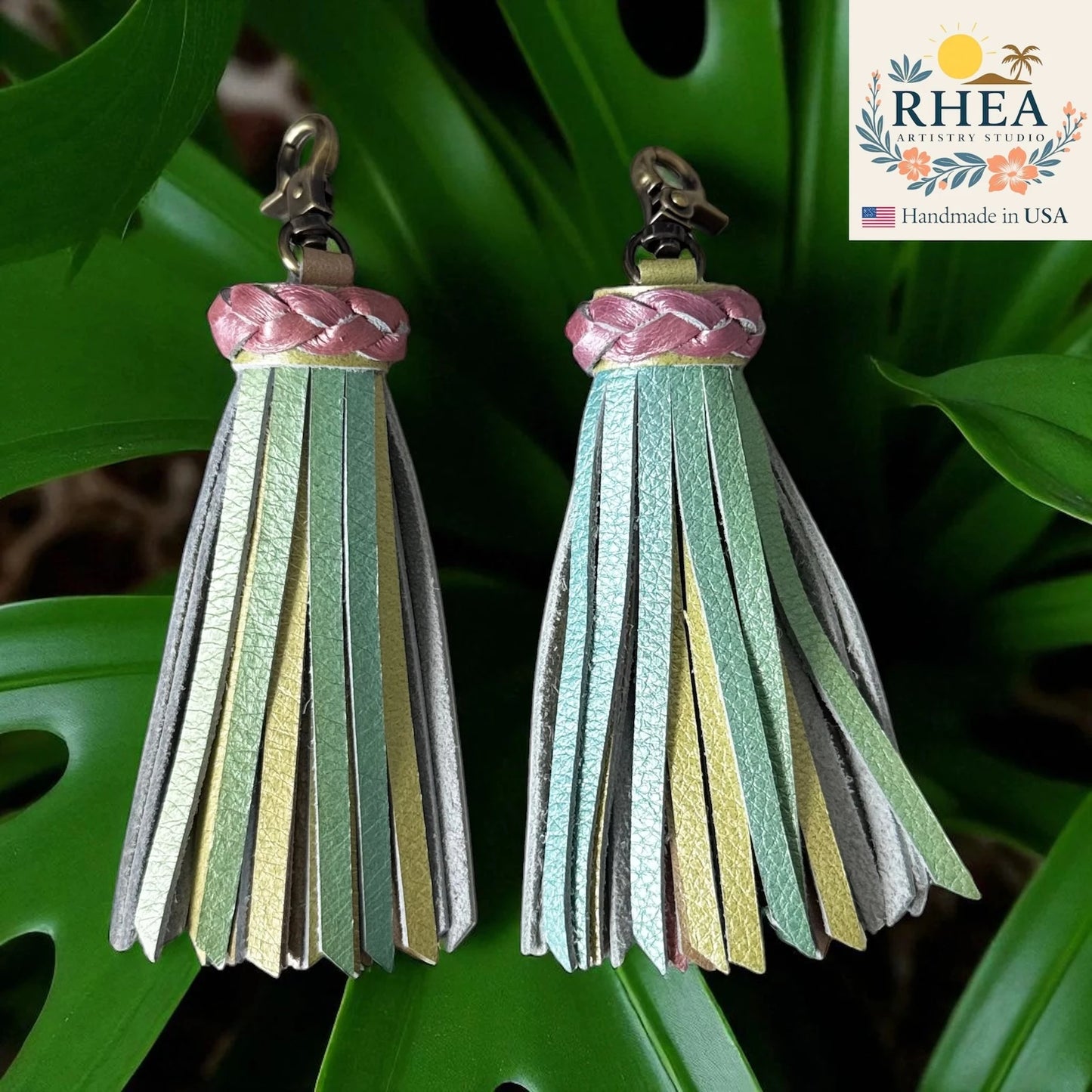 Pair of leather tassels with braided top on a green leaf background, featuring the RHEA brand.