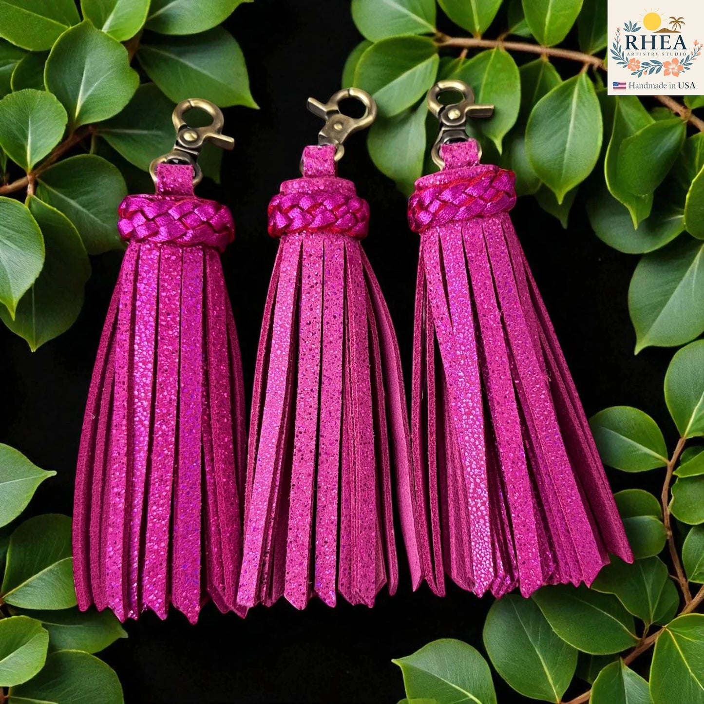 Three hot pink tassel keychains on a green leaf background with RHEEA branding.