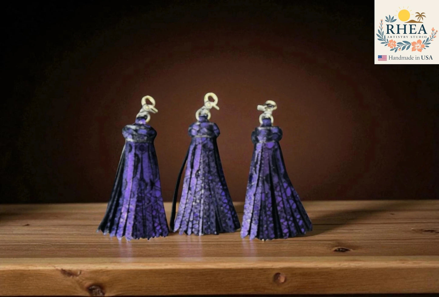 Three purple tassels on a wooden surface with a dark background, featuring the RHEA brand logo.