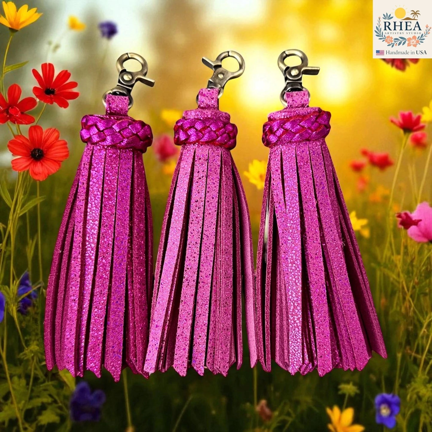 Three hot pink tassels with metal clasps against a colorful floral background, featuring the brand 'RHEA'.