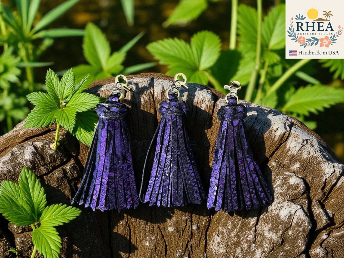 Three purple tassel earrings on a wooden block with green leaves in the background, featuring the RHEA brand.