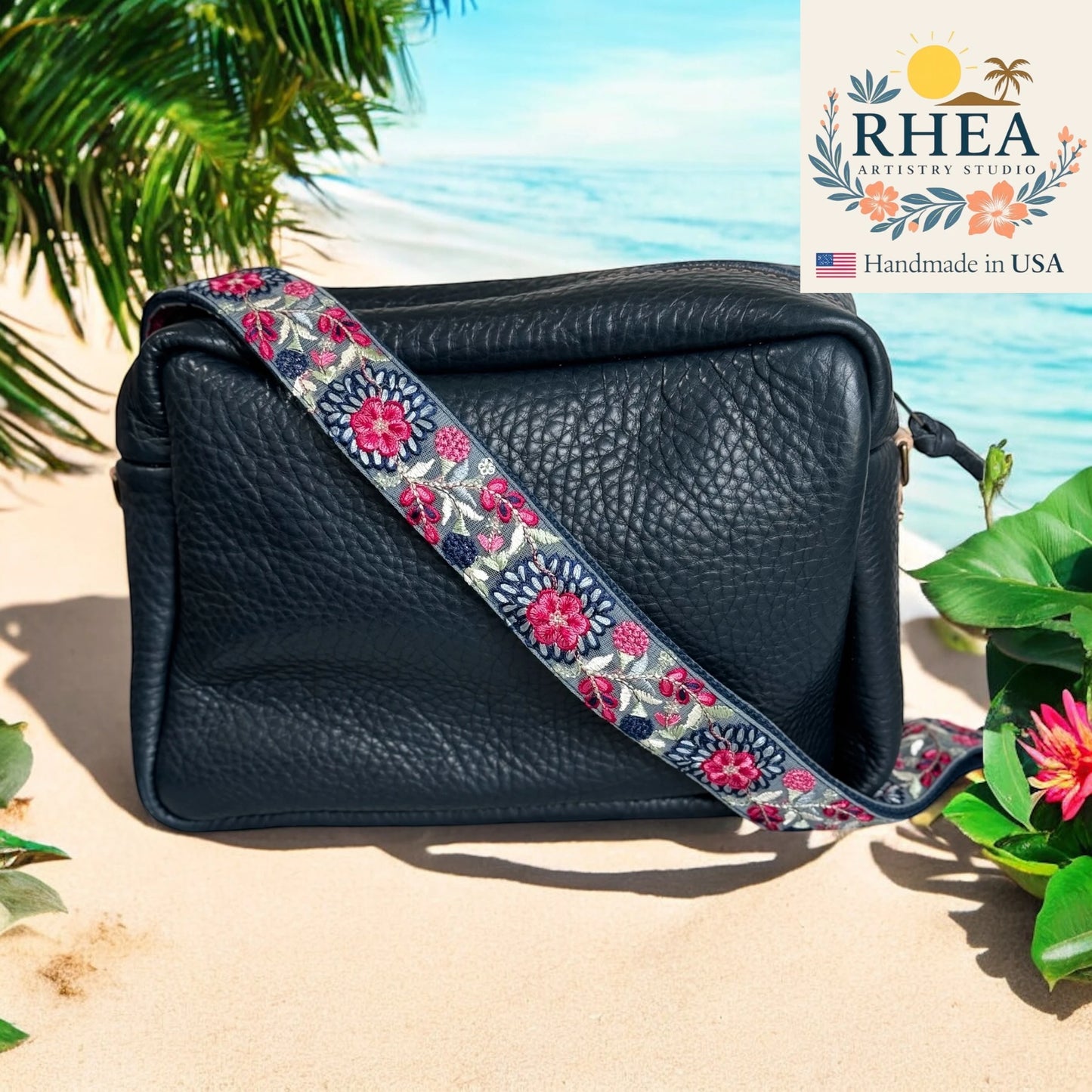 Black leather handbag with a floral purse strap on a beach background, featuring the RHEA Artistry Studio logo.