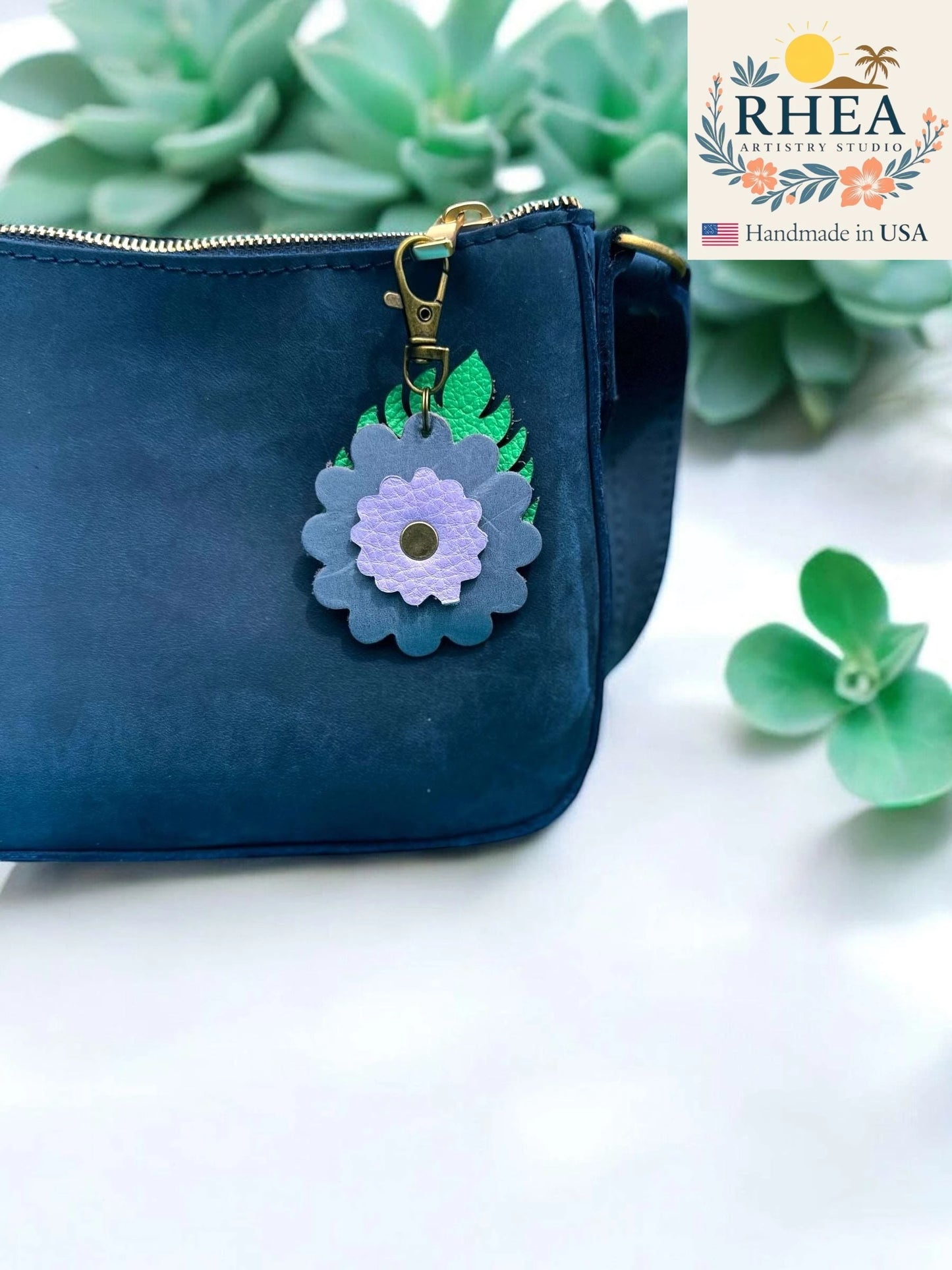 Blue pouch with a purse charm on a white surface with succulents, featuring the RHEA brand.
