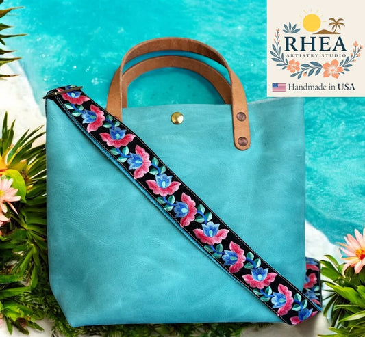 Turquoise tote bag with floral strap against a blue background, featuring the Rhea Artistry Studio logo.