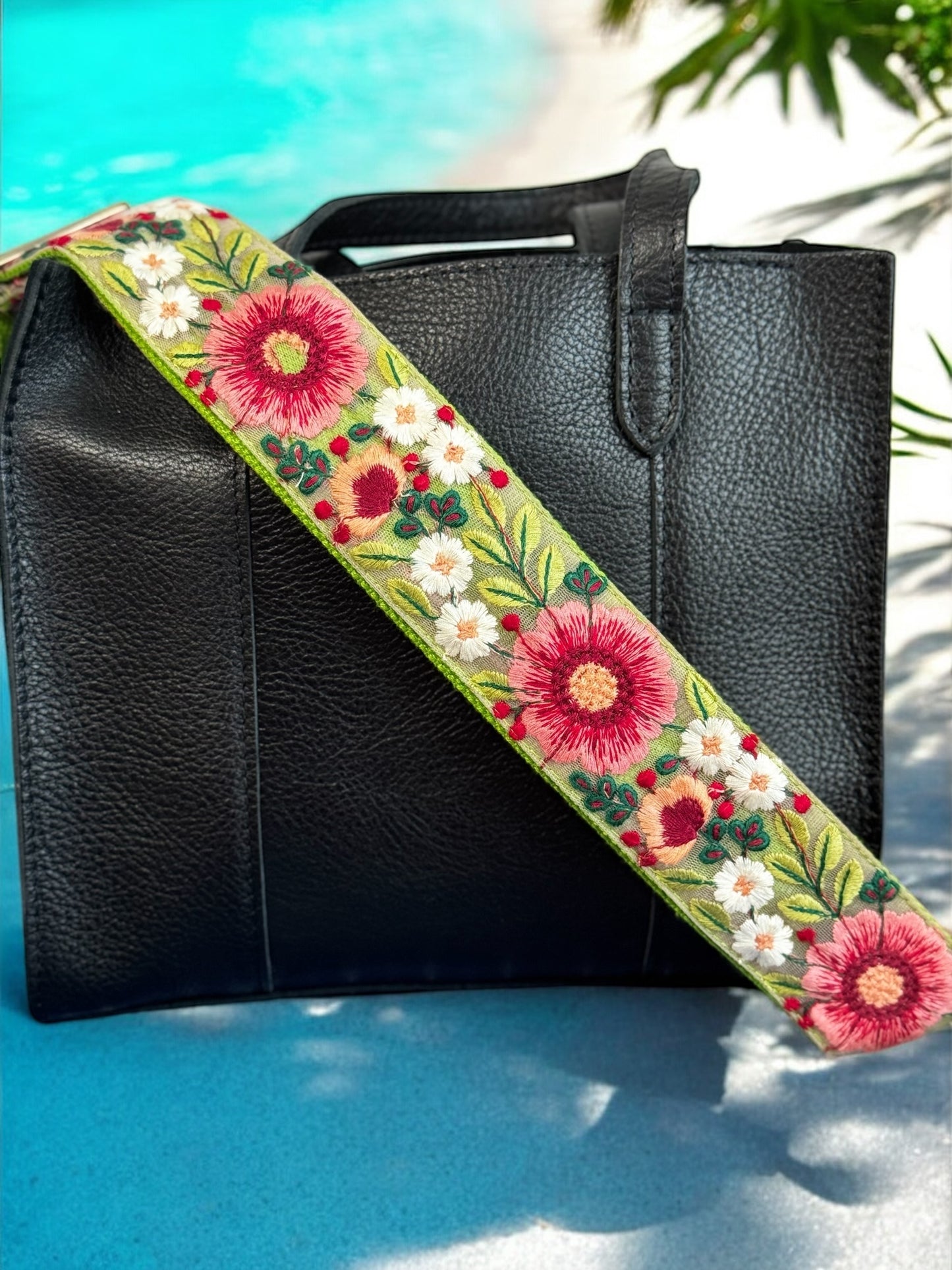 Olive green floral crossbody strap with crimson daisies and ivory blossoms attached to a structured black leather handbag at beach