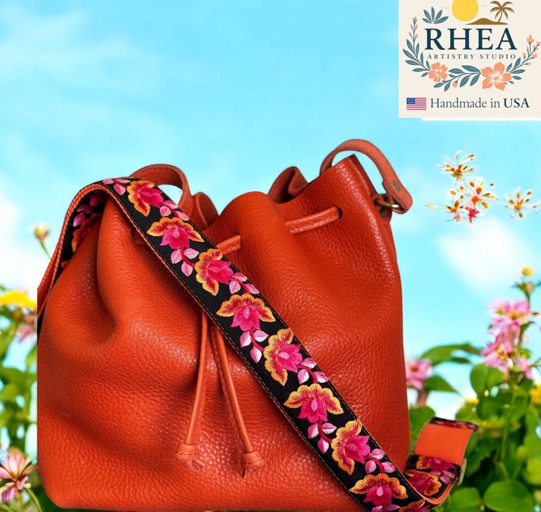 Orange leather bag with a floral purse strap against a blue sky and greenery background, featuring the RHEA Artistry Studio logo.