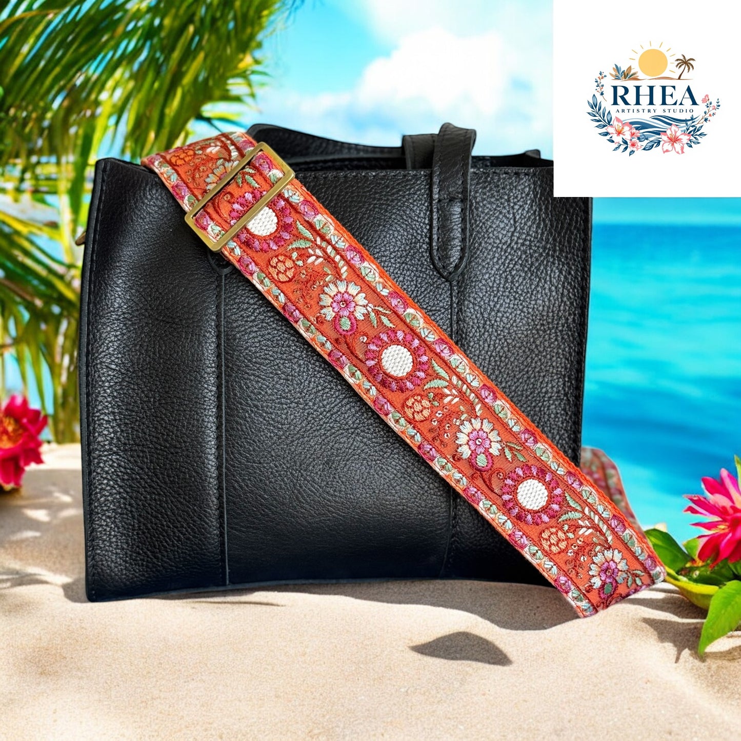 A bold marigold-orange embroidered strap featuring intricate floral motifs in shades of pink, white, and sage green, paired with a structured black bag against a sandy beach
