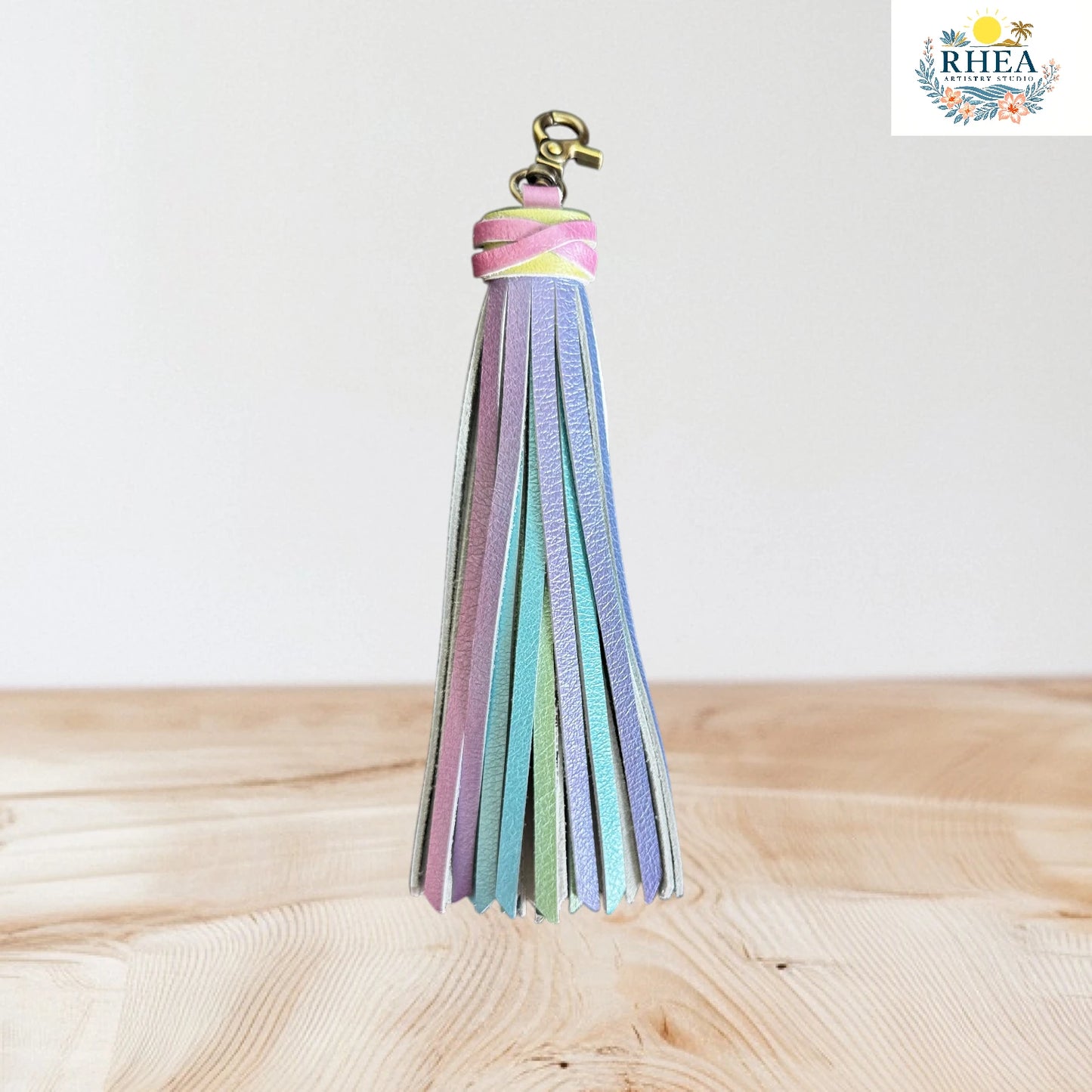 Colorful tassel keychain on a wooden surface with 'RHEA' branding in the corner.