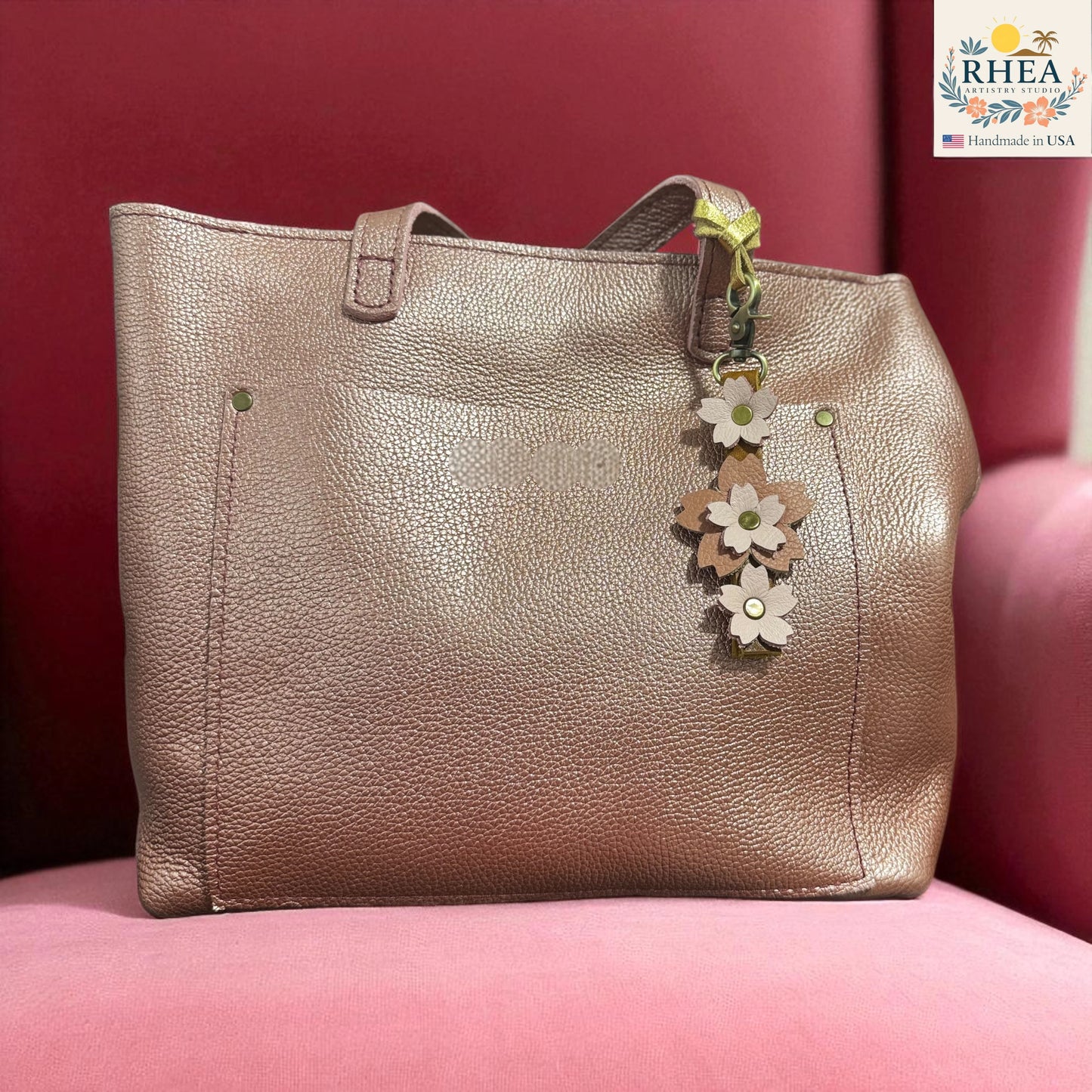 Gold handbag with a floral charm on a pink surface, featuring the RHEA brand.