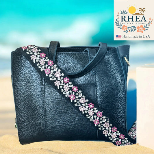 Black leather handbag with floral strap on a beach background, featuring the RHEA Artistry Studio logo.