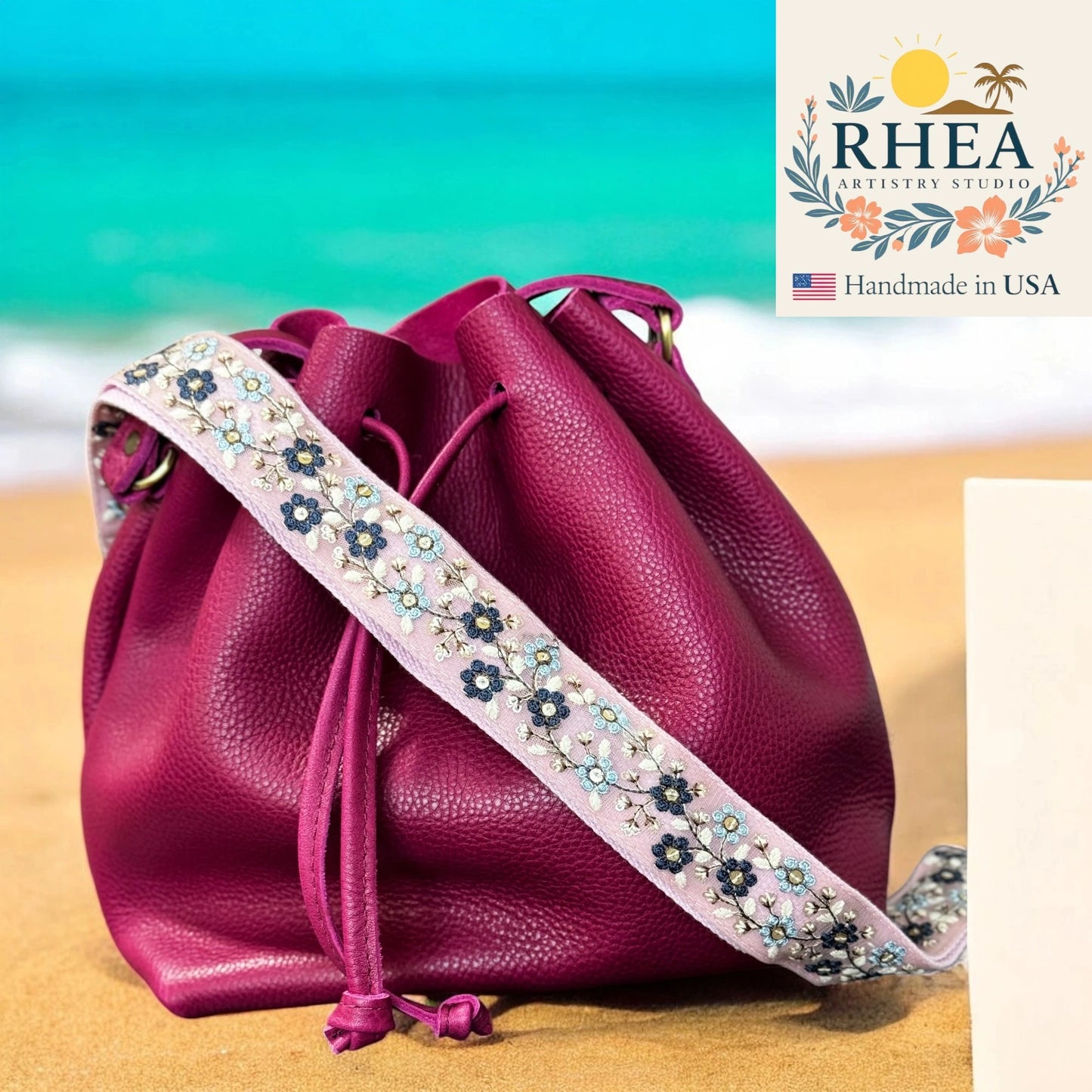 Pink leather drawstring bag with floral strap on a beach background, featuring the RHEA brand.