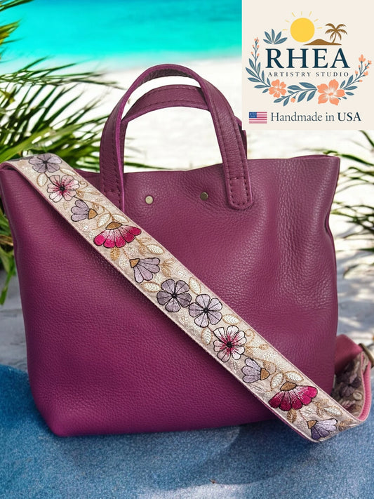 Purple handbag with a floral purse strap on a blue surface, branded 'RHEA Artistry Studio'.
