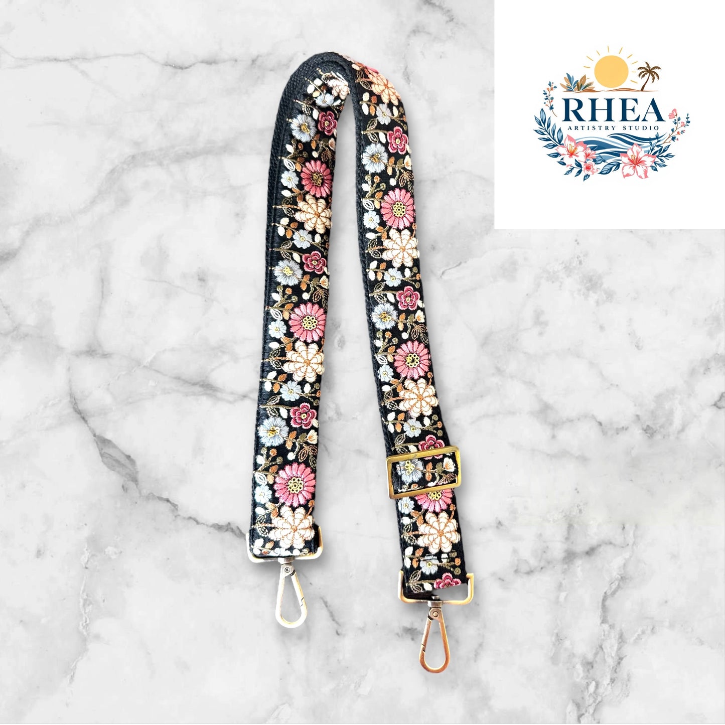 An adjustable strap with daisy flowers embroidery attached to a black bag, with a field and sunset in the background.