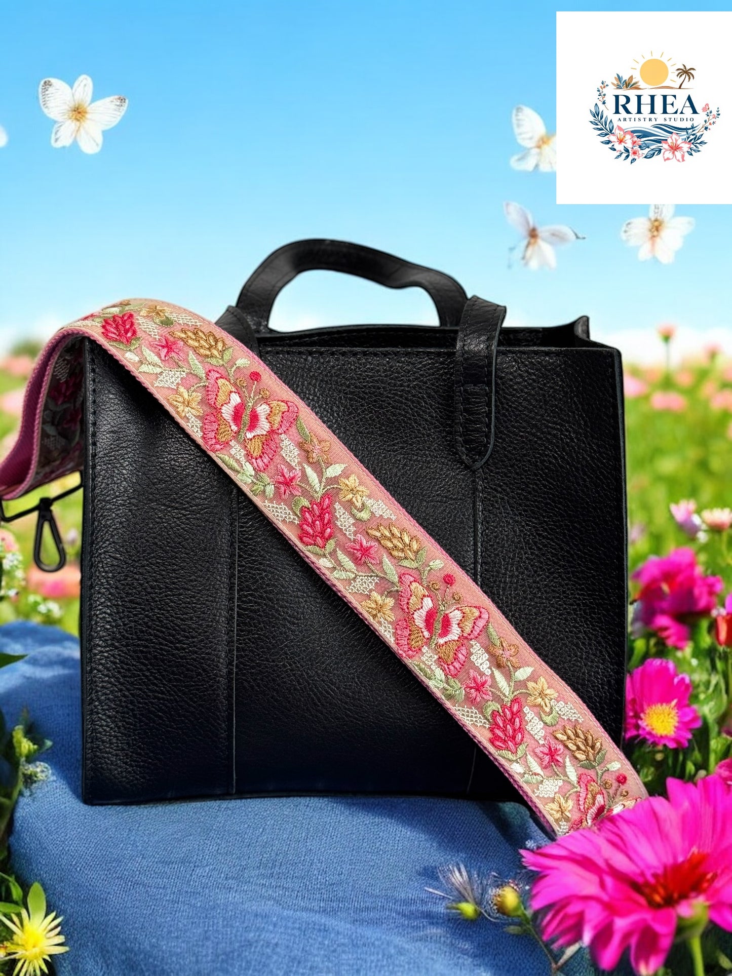 A black purse with a pink and green floral embroidered strap placed on a blue surface with flowers in the background.