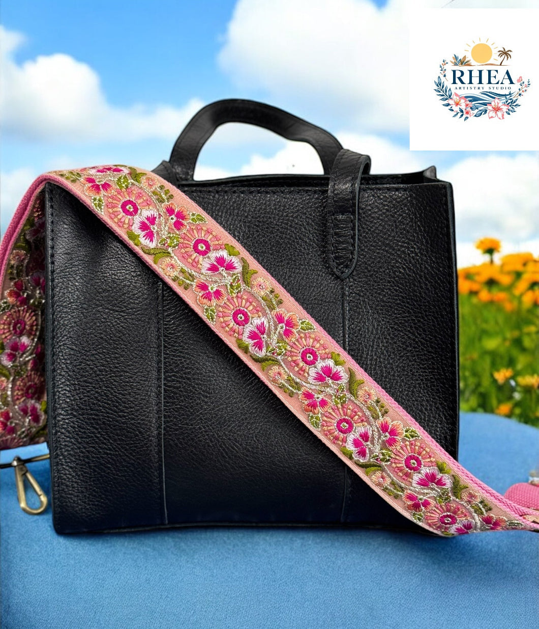 A black purse with a pink and green floral embroidered strap placed on a blue surface with a bright sky and greenery in the background.