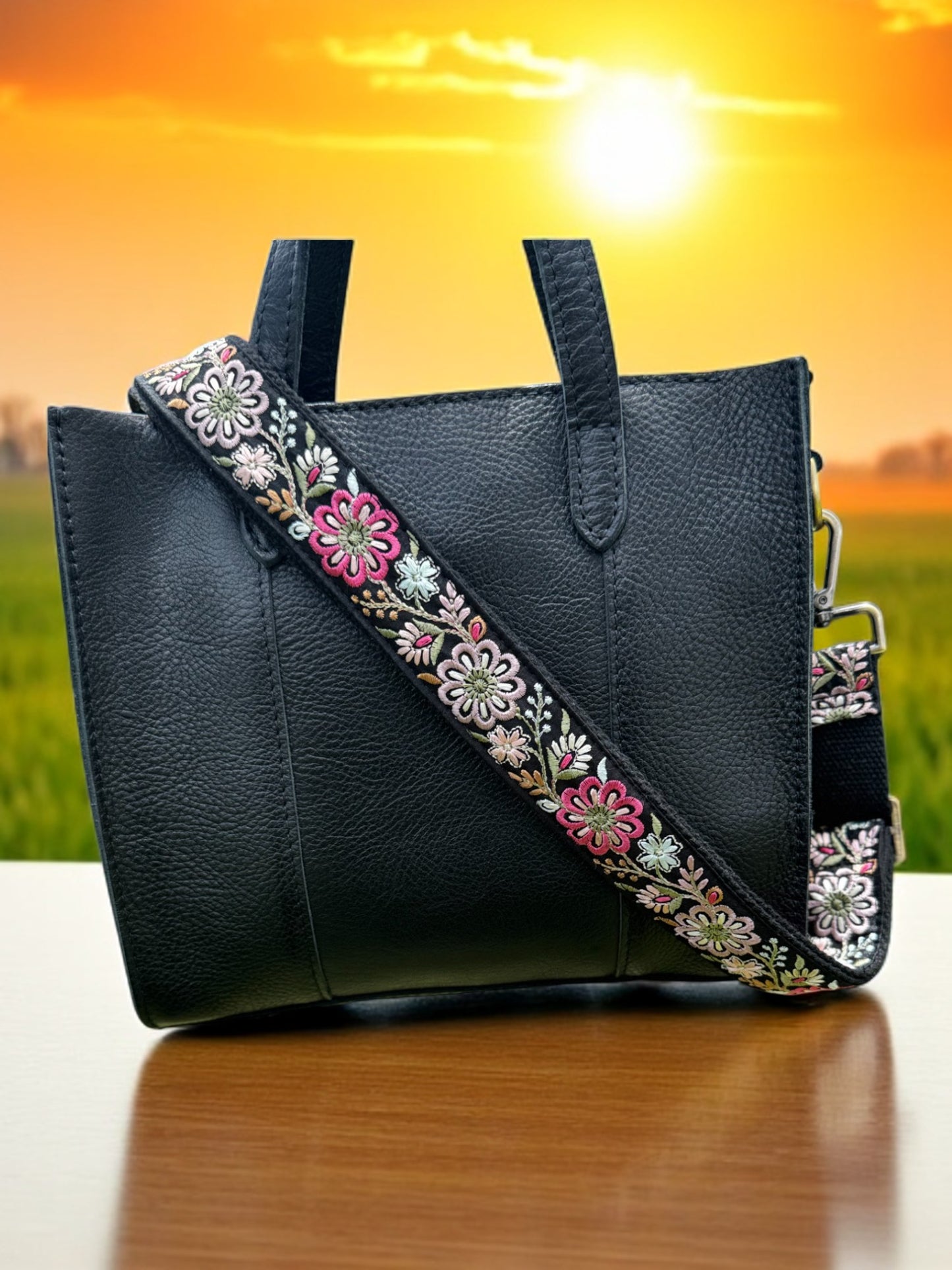 Black bag with vibrant floral crossbody strap featuring pink, ivory, and olive green tones against sunset background.