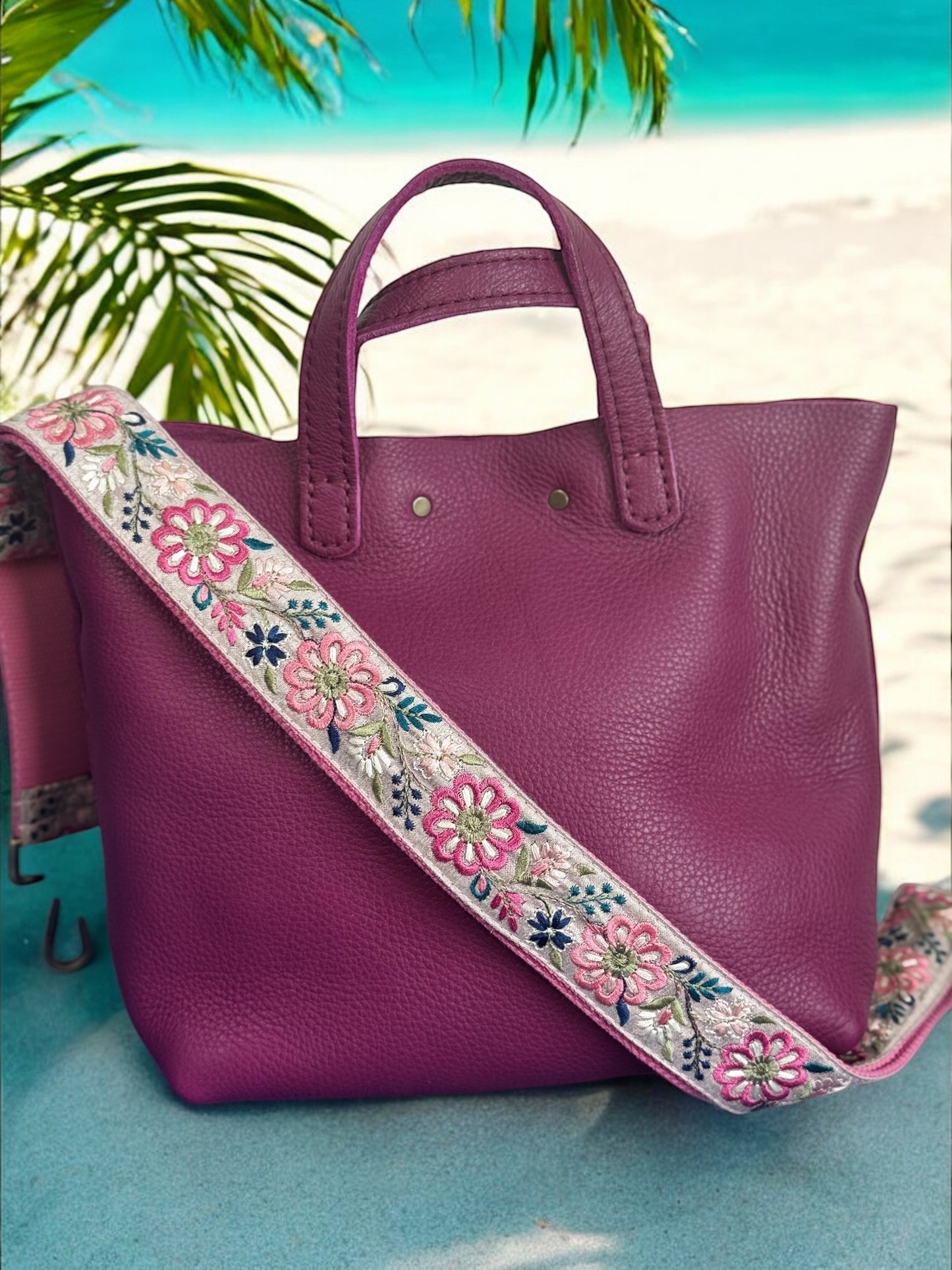 Magenta handbag with vibrant pink and white floral strap in pinwheel embroidery set against a beach backdrop.