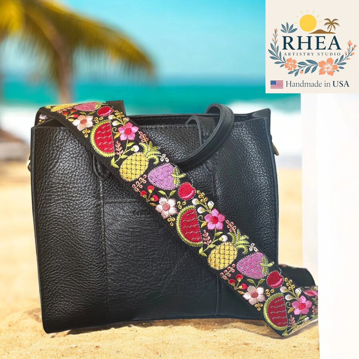 Black handbag with colorful floral strap on a beach background, featuring RHEA Artistry Studio logo.