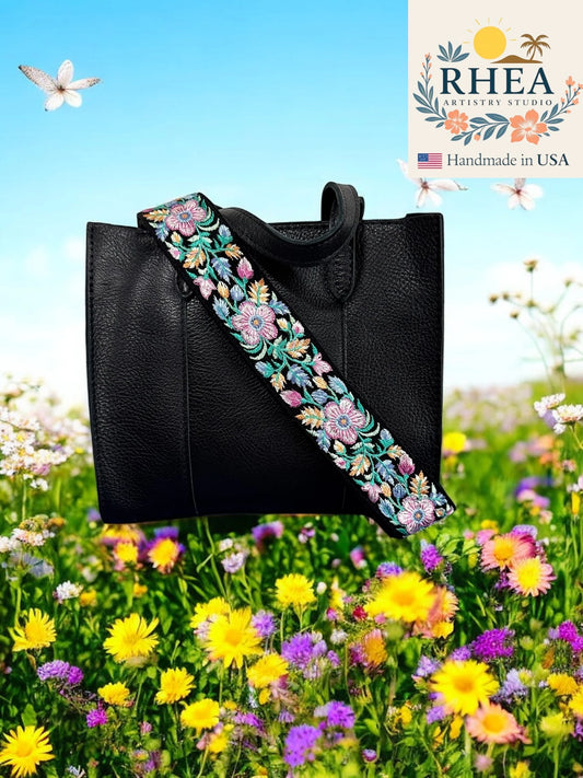 Black handbag with a floral purse strap in a field of flowers, featuring the RHEA Artistry Studio logo.