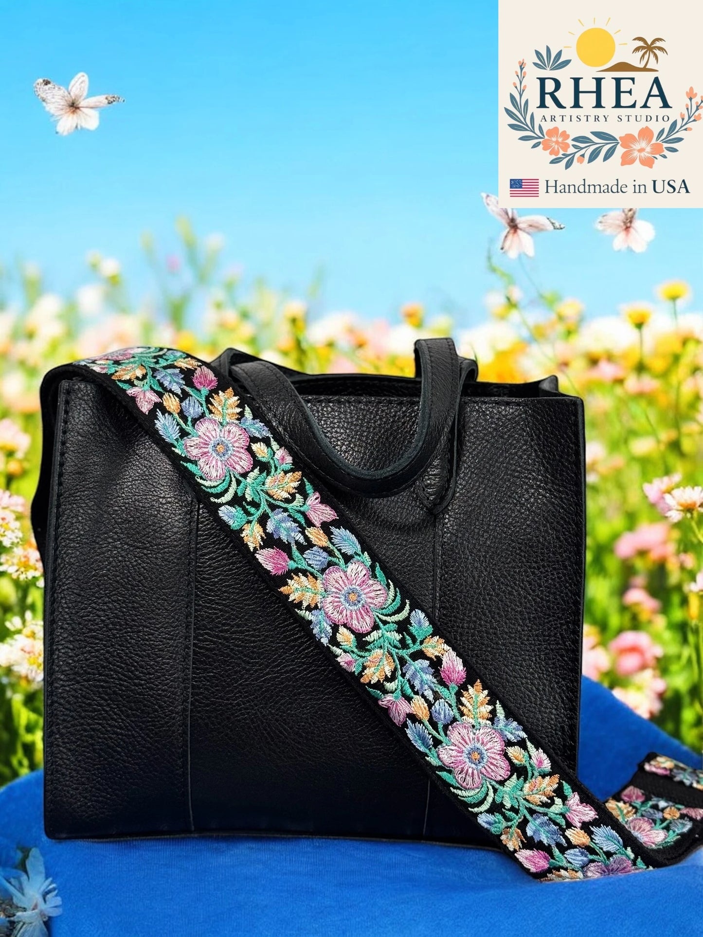 Black handbag with a floral purse strap against a blue sky and flowers background, featuring the Rhea Artistry Studio logo.