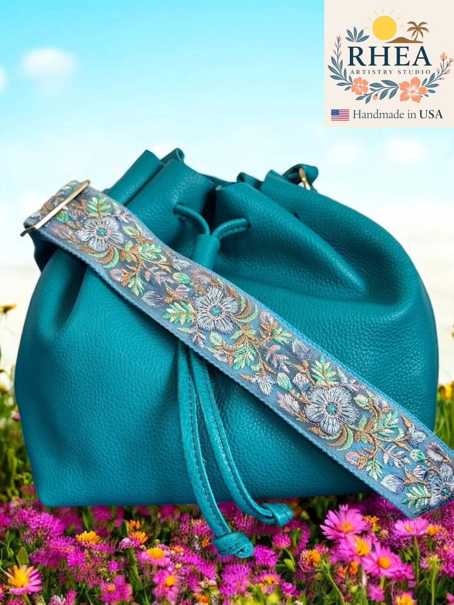 Teal bag with a floral purse strap in a field of flowers, featuring RHEA Artistry Studio branding.