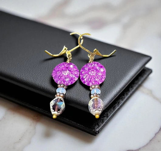 Handcrafted dangle earrings with vibrant purple floral glass discs, iridescent beads, and gold-plated hooks, displayed on a black leather cushion.