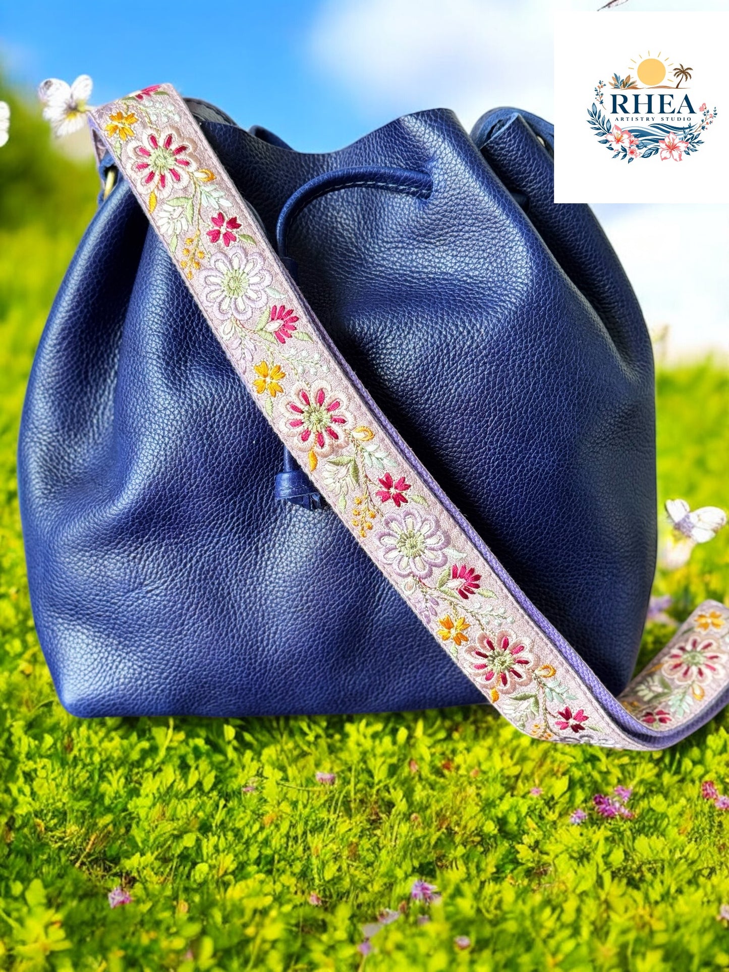 Adjustable handmade floral strap in a whimsical pinwheel design on a lilac background, paired with a navy pebbled leather bag, captured in a meadow.