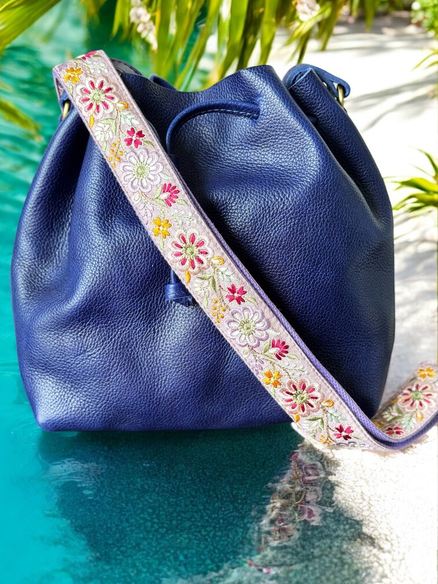 Adjustable handmade floral strap in a whimsical pinwheel design on a lilac background, paired with a navy pebbled leather bag, captured in a beach.