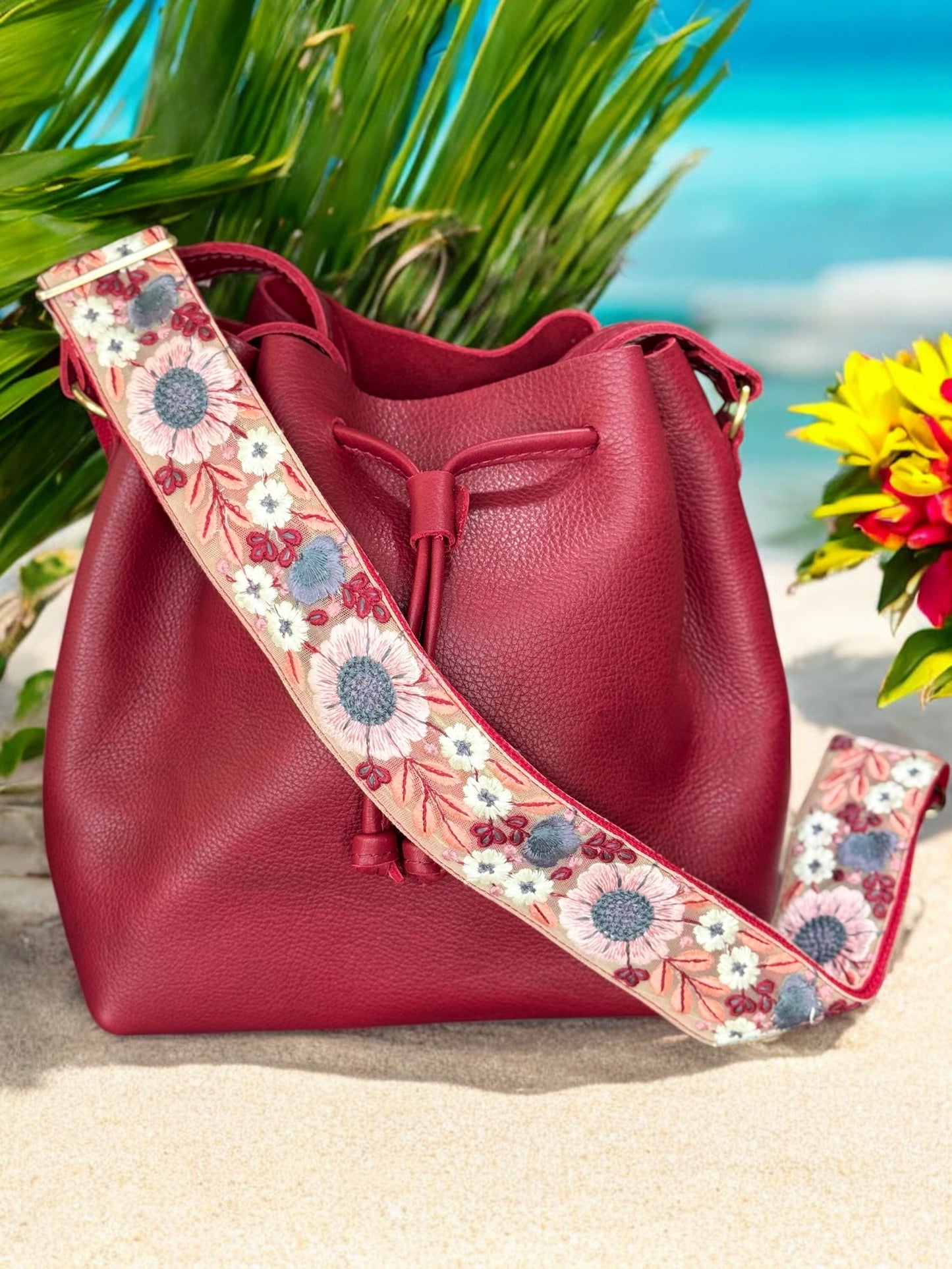 Handcrafted red crossbody bag strap with intricate floral embroidery in pink, blue, and white, shown attached to a matching red bucket bag on a sandy beach with palm leaves in the background. Made in USA.