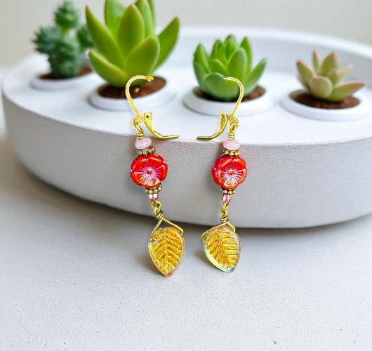Pair of earrings with red and gold leaf designs on a white surface with succulents in the background.