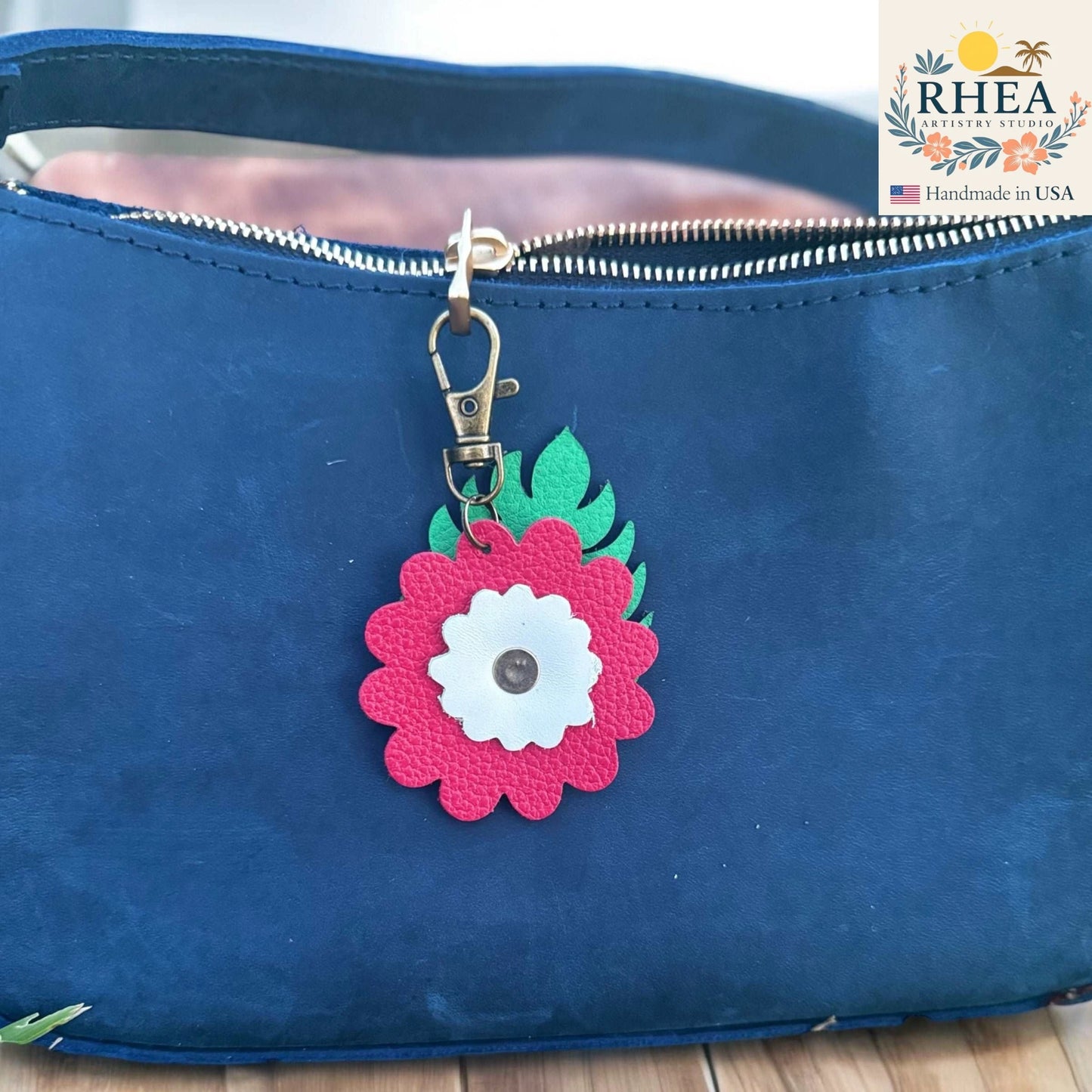Blue bag with a pink floral bag charm on a wooden surface, featuring the RHEA brand.