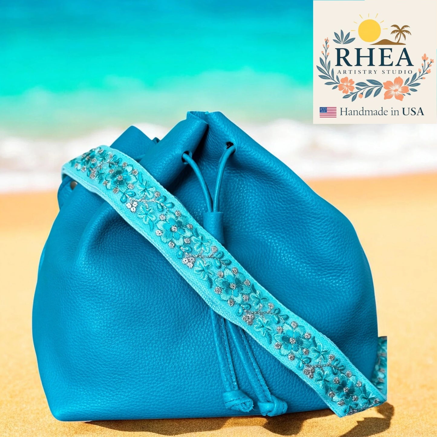 Blue handbag with floral embroidery on a beach background, featuring the RHEA Artisan Studio logo.