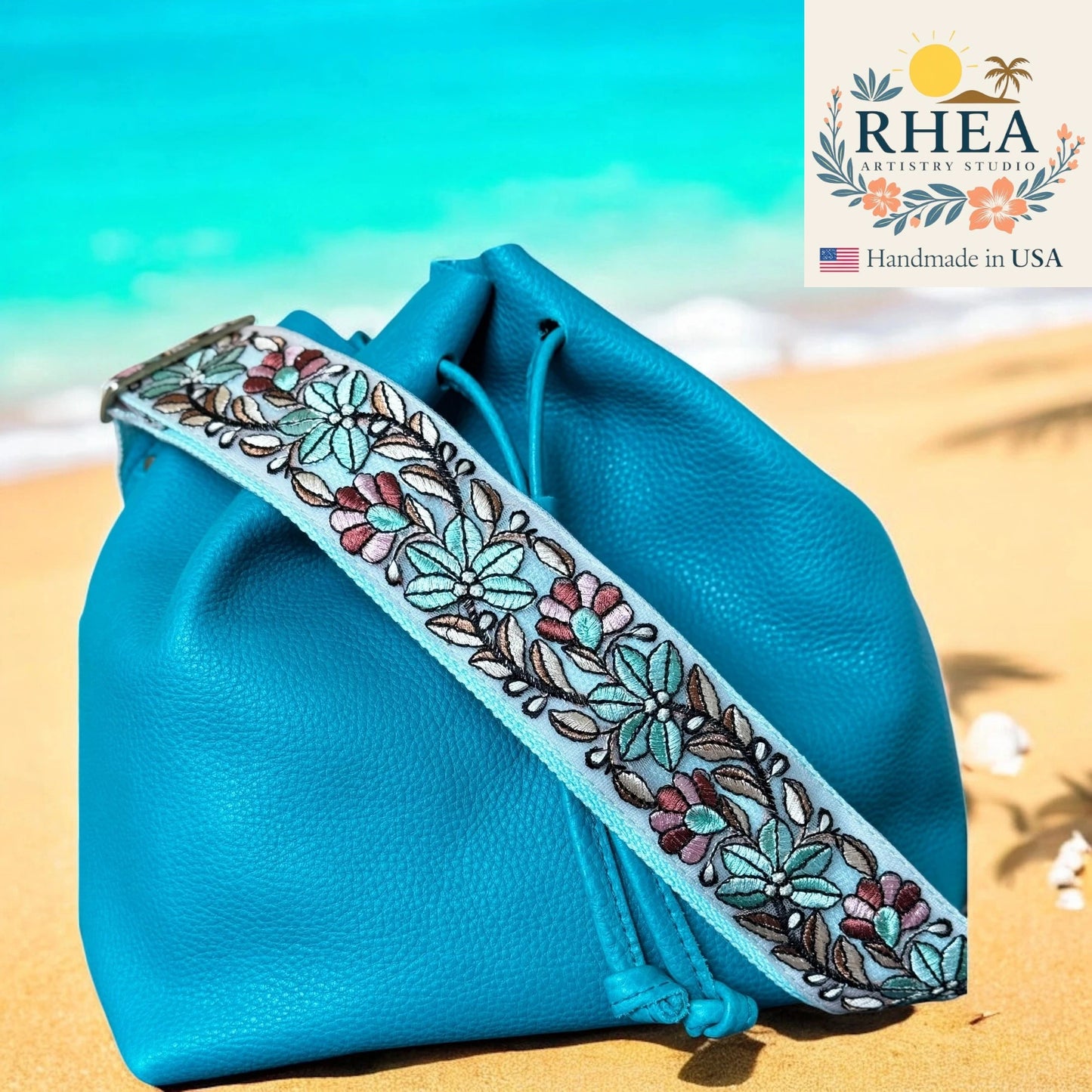 Blue pouch with floral embroidery purse strap on a beach background, featuring the RHEA Artistry Studio logo.