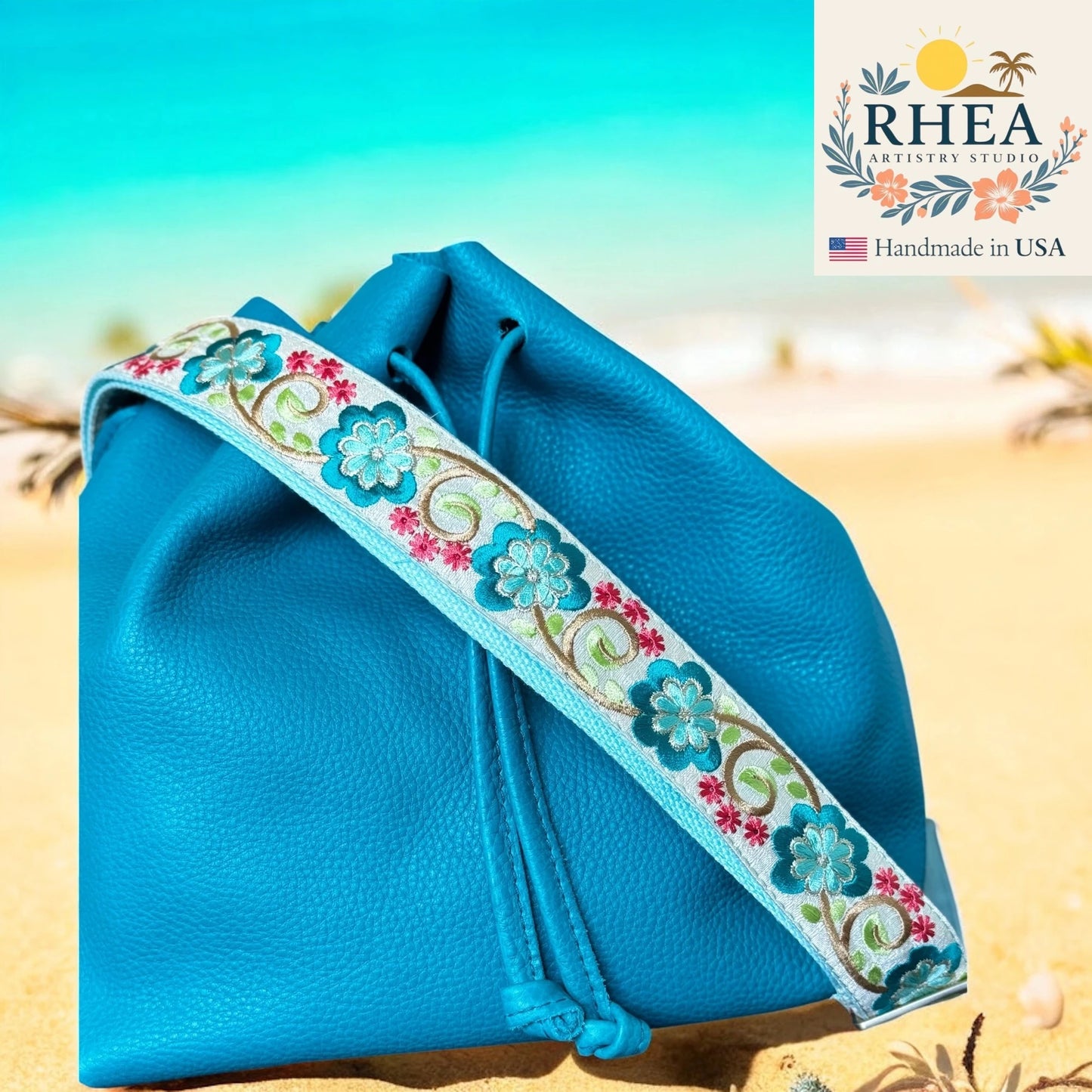 Blue drawstring bag with floral purse strap on a beach background, featuring the RHEA brand.