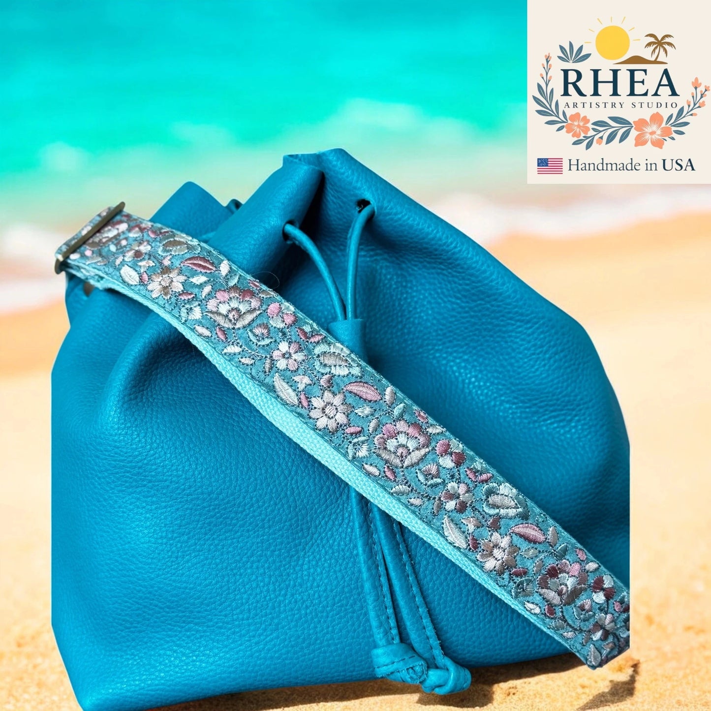 Blue drawstring bag with floral purse strap on a beach, featuring RHEA Artistry Studio logo.
