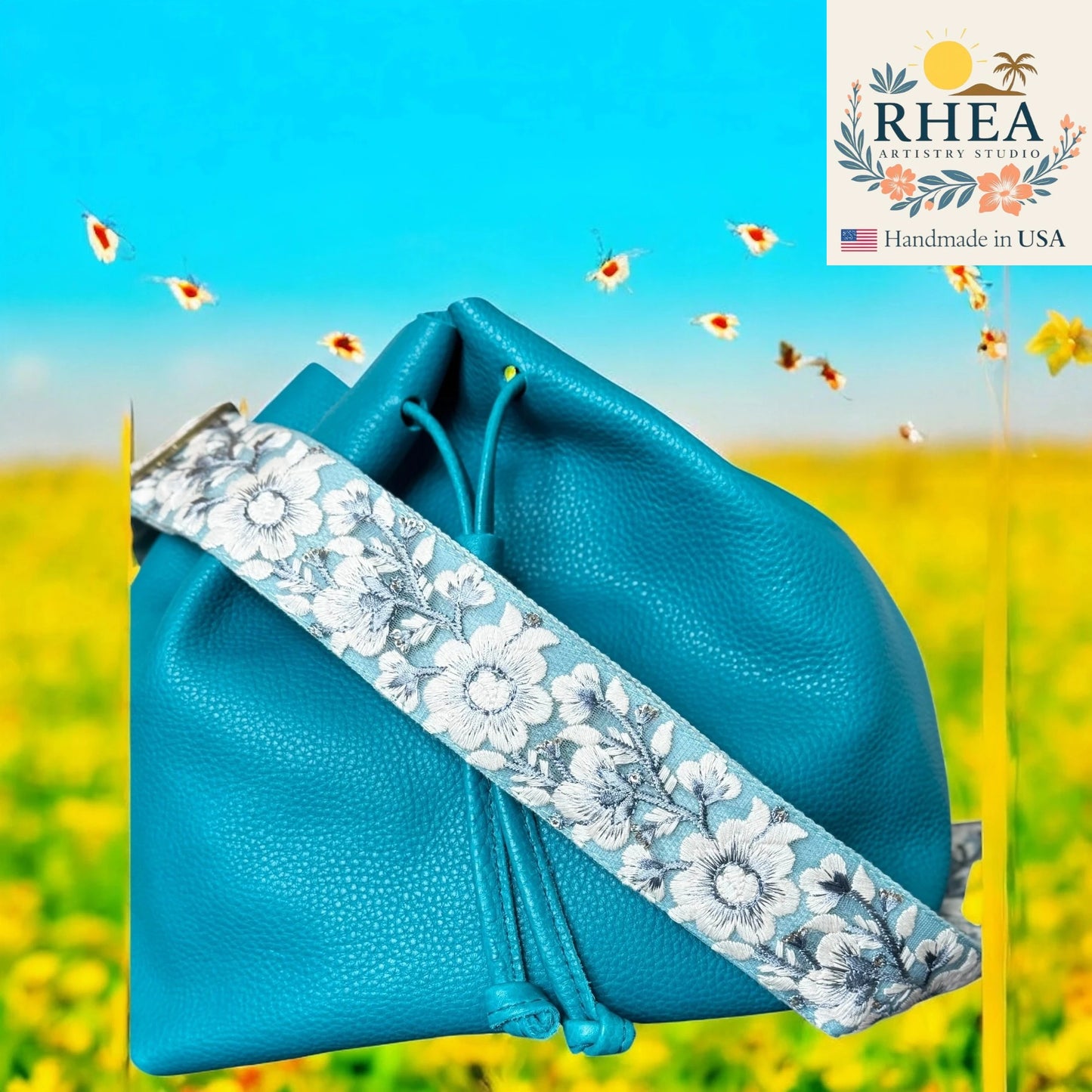 Blue pouch with floral strap against a blurred natural background, featuring the RHEA brand.