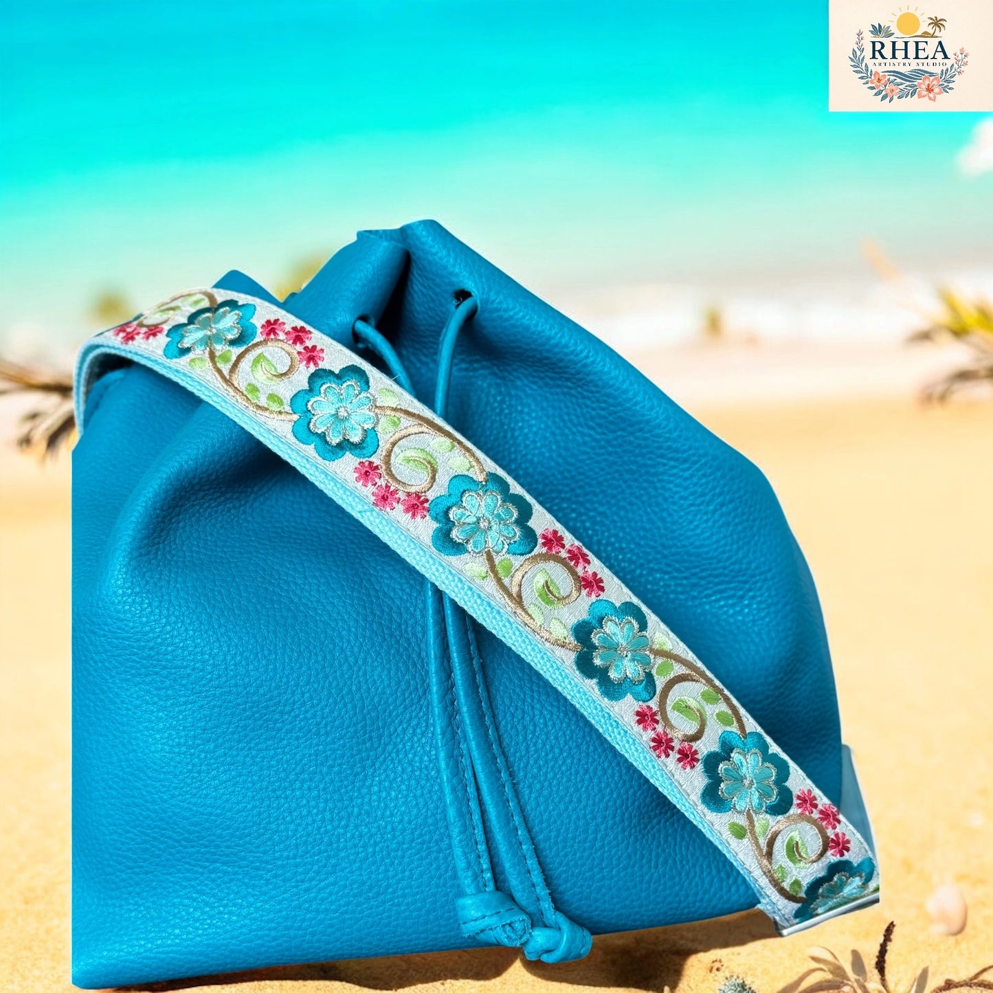 Blue drawstring bag with floral strap on a beach background, featuring the RHEA brand.