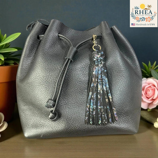 Rhea Artistry Studio black leather tassel keychain with glitter accents and antique brass lobster clasp attached to a black drawstring bag.