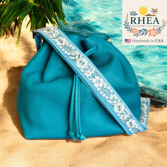 Rhea Artistry Studio blue crossbody strap with white floral embroidery, 1.5-inch wide, adjustable length, on a teal bucket bag.