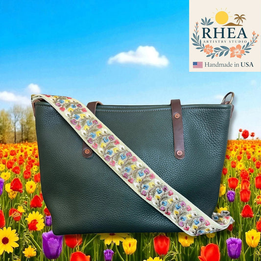 Rhea Artistry Studio 2-inch wide crossbody strap with embroidered floral lace in blue, pink, and yellow on a cream background, attached to a dark green pebbled leather tote bag.