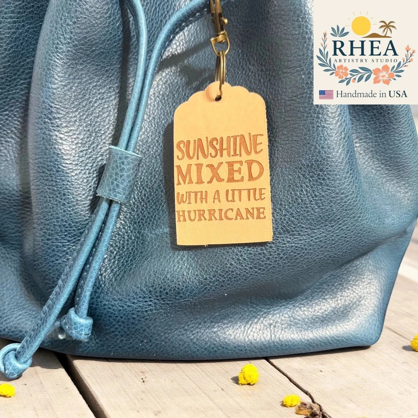 Rhea Artistry Studio natural leather tag engraved with "Sunshine Mixed With A Little Hurricane" hanging from a blue leather bag.