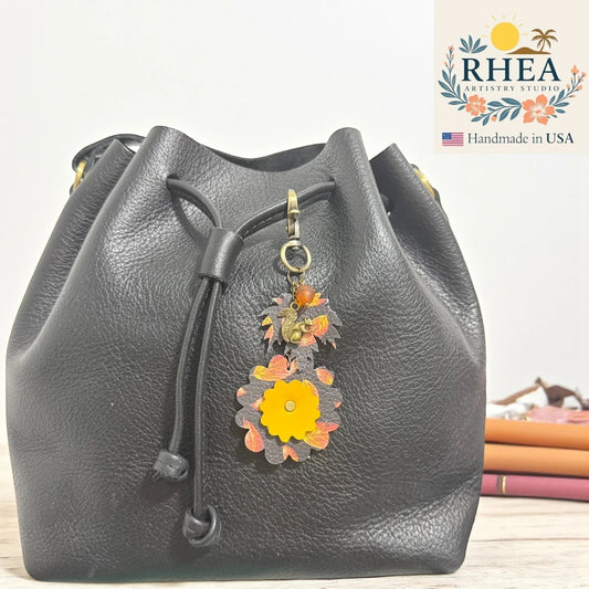 Rhea Artistry Studio fall leather charm with acorn and squirrel silhouettes, autumn leaves, and a yellow flower, attached to a black leather bag.