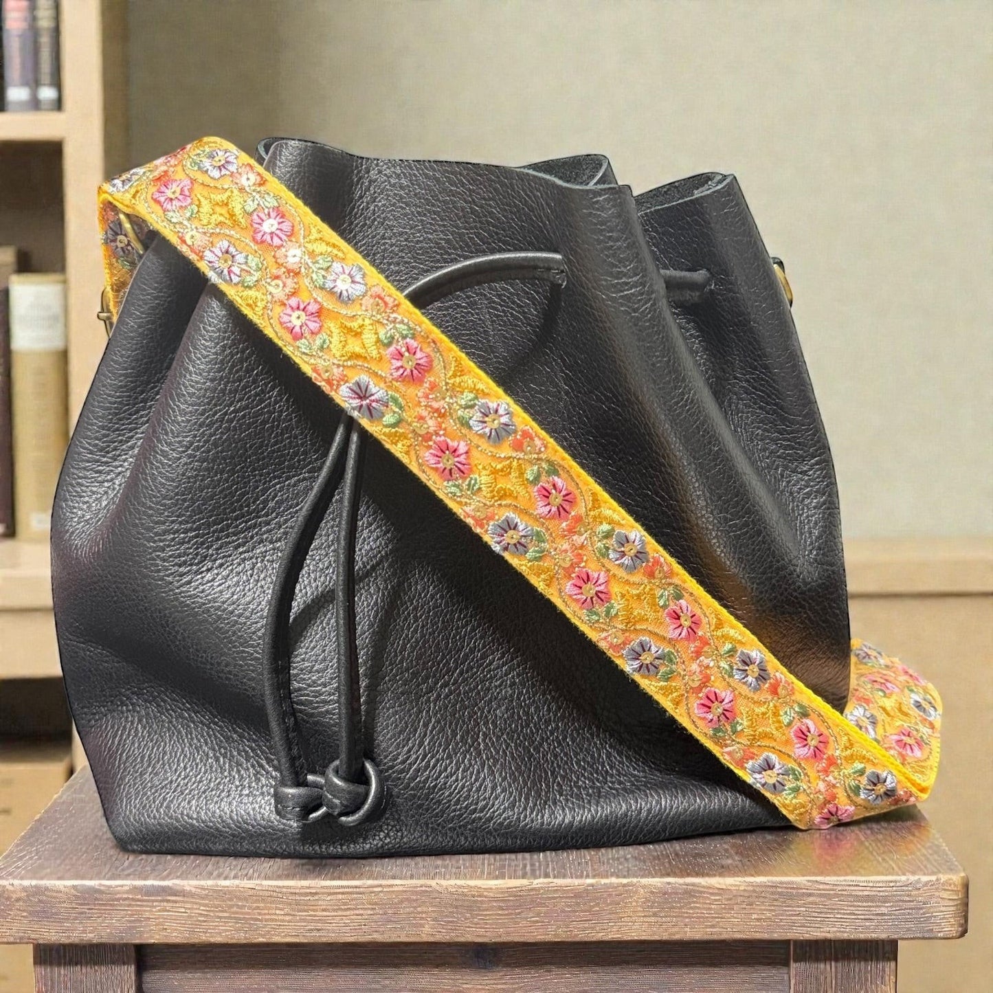 Rhea Artistry Studio yellow crossbody strap with floral embroidery and brushed brass hardware on a black leather bucket bag.