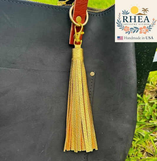 Rhea Artistry Studio jumbo gold full-grain leather tassel keychain with lobster clasp attached to a black handbag.