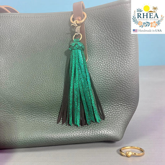 Rhea Artistry Studio handmade green leather tassel keychain with lobster clasp attached to a grey pebbled leather tote bag.