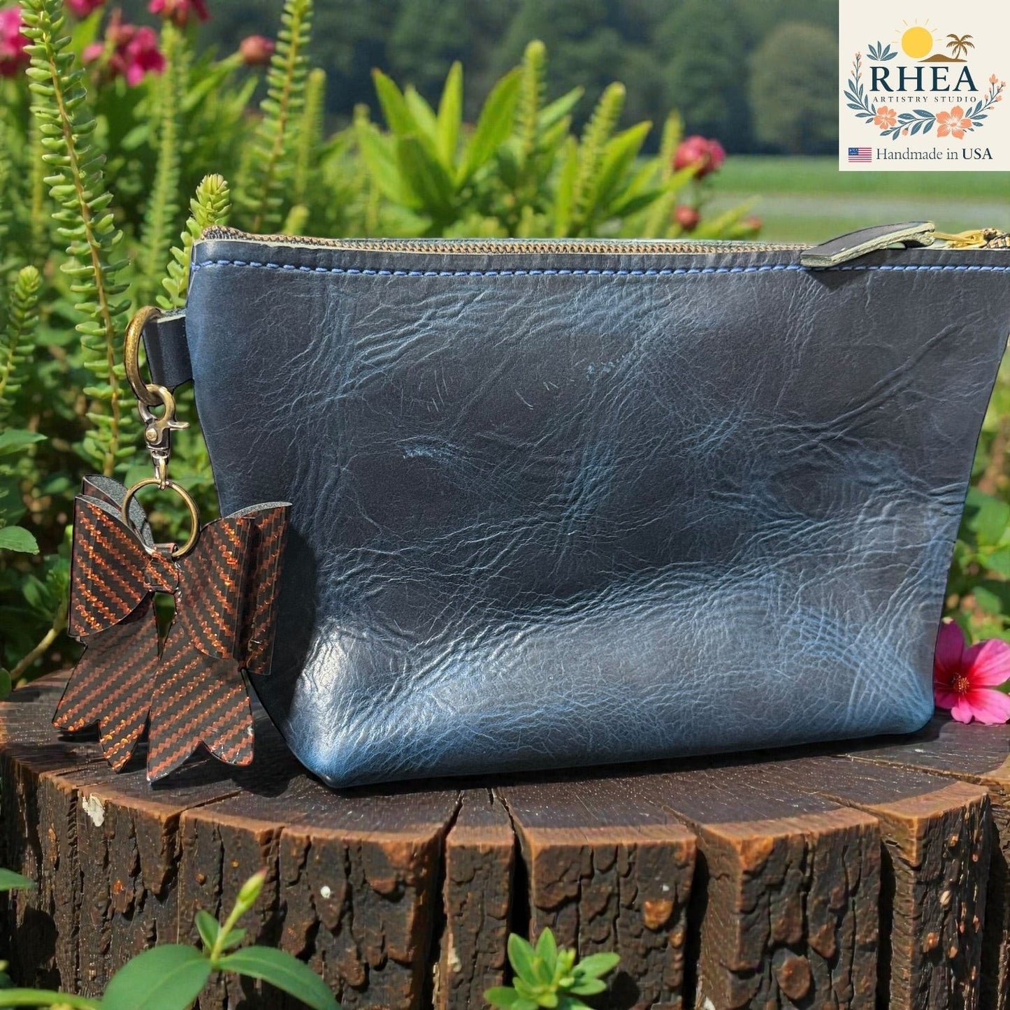 Rhea Artistry Studio holographic bow bag charm with carbon fiber pattern and brass hardware, attached to a blue leather pouch.