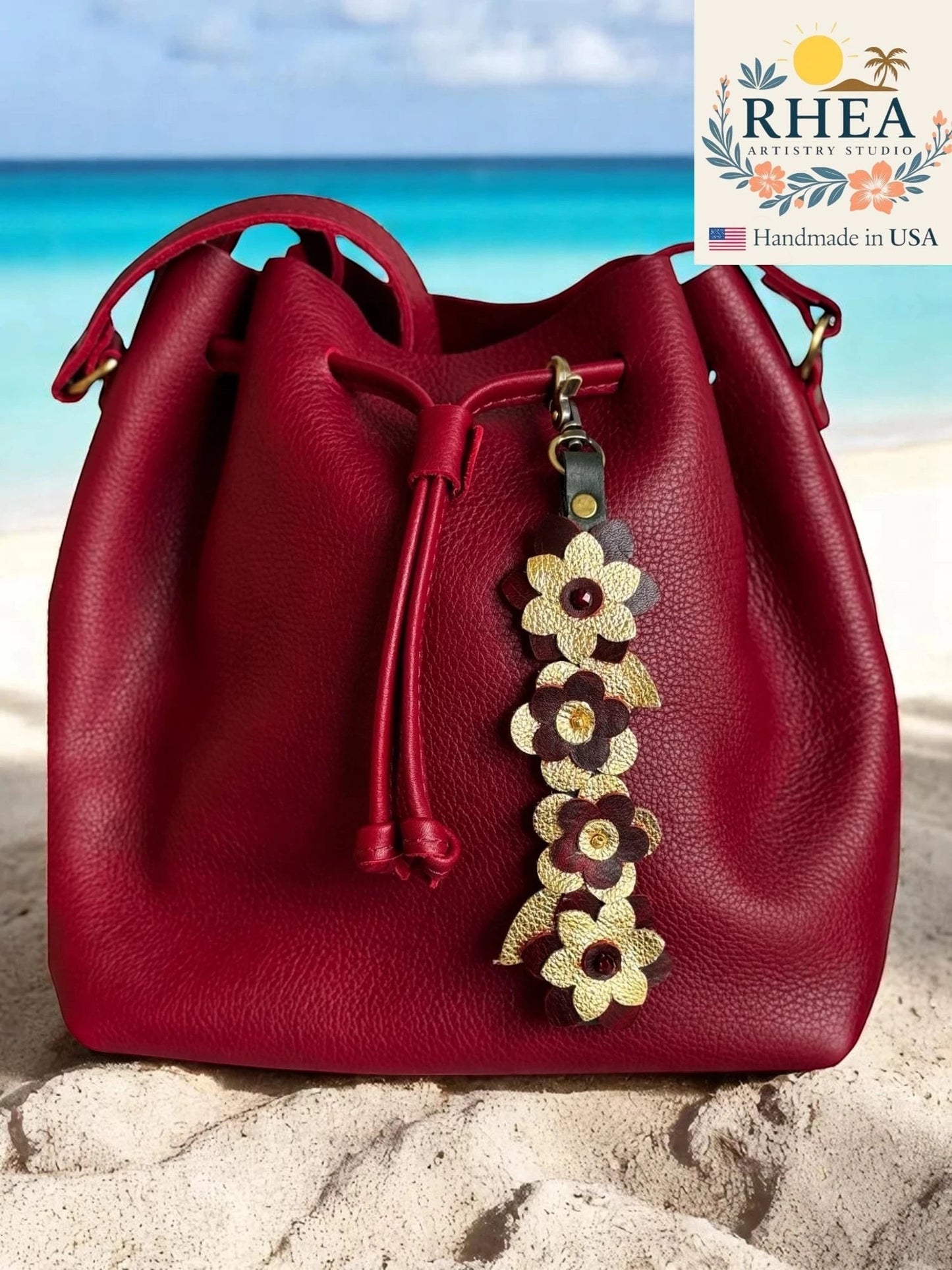 Rhea Artistry Studio handcrafted leather charm with gold and maroon flowers and Czech beads, attached to a red leather bucket bag on a sandy beach.
