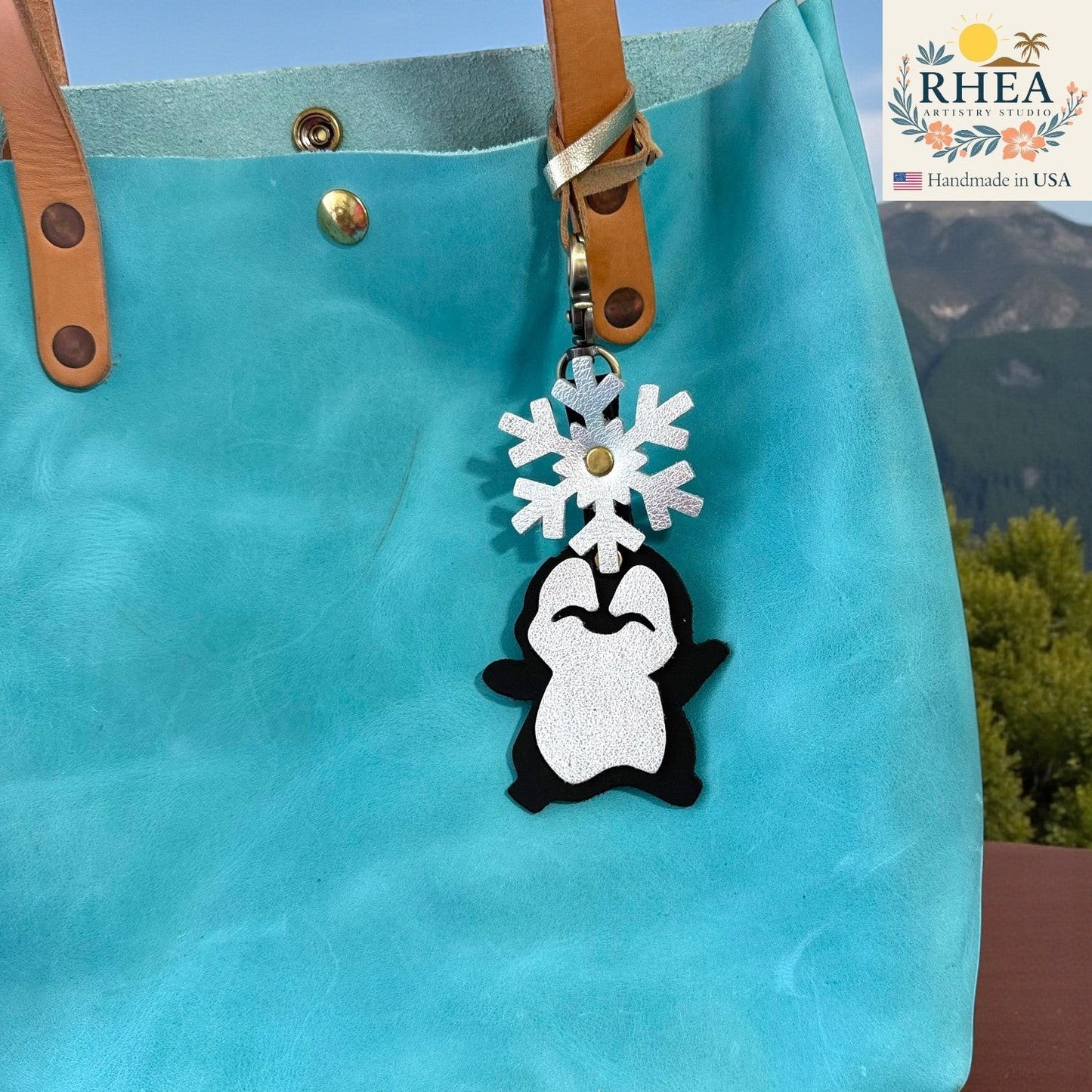 Rhea Artistry Studio handmade black and white leather penguin charm with a silver snowflake detail, attached to a teal tote bag.